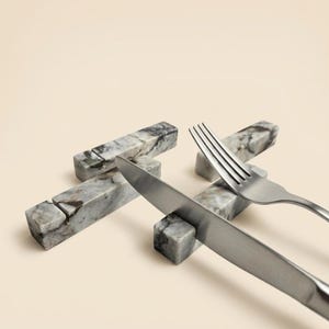 May include: A set of three rectangular grey and white marble chopstick or cutlery rests, each featuring a distinct veined pattern and a central groove. A stainless steel knife and fork are positioned resting across two of the marble pieces.