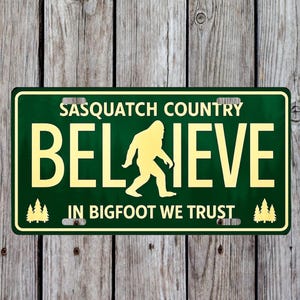 May include: A green license plate with the words "SASQUATCH COUNTRY" above the word "BELIEVE" and a Bigfoot silhouette. The phrase "IN BIGFOOT WE TRUST" is below. The plate has a yellow border and text.