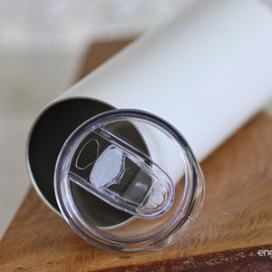 May include: A white insulated tumbler with a clear plastic lid. The lid is partially open, revealing the interior of the tumbler. The tumbler is cylindrical and sits on a wooden surface. The tumbler is designed to keep beverages hot or cold.
