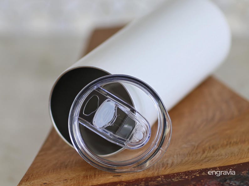 May include: A white insulated tumbler with a clear plastic lid. The lid is partially open, revealing the interior of the tumbler. The tumbler is cylindrical and sits on a wooden surface. The tumbler is designed to keep beverages hot or cold.