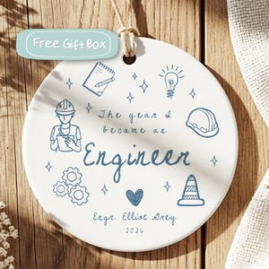 Puede incluir: Un adorno redondo de cerámica blanca con ilustraciones de línea azul de una persona con casco, un bloc de notas, una bombilla, engranajes y un cono de tráfico. El texto dice: The year I became an Engineer, Engr. Elliot Grey, 2026.