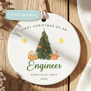 May include: A round white ceramic ornament featuring a watercolor Christmas tree, a toolbox, and a hard hat. The text reads "FIRST CHRISTMAS AS AN ENGINEER", "ENGR. ELLIOT GREY", and "2026". It hangs from a jute twine loop at the top center.