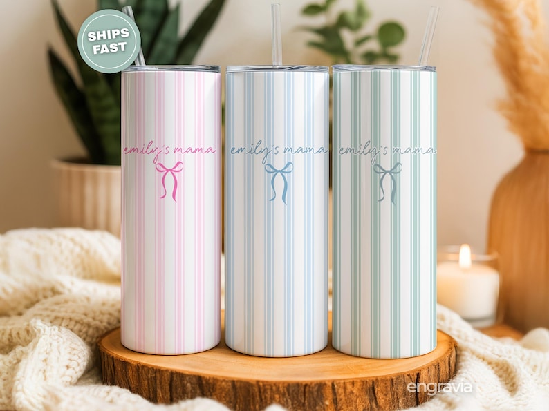 May include: Three striped tumblers in pink, blue, and green, each with a bow graphic and the text "emily's mama". The tumblers are displayed on a wooden surface, with a straw in each. The background includes a plant and a candle.