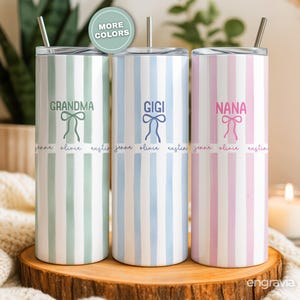 May include: Three personalized stainless steel tumblers with vertical stripes in pastel colors: green, blue, and pink. Each tumbler has a name: "Grandma," "Gigi," and "Nana," with a bow graphic and names below. The tumblers have a silver straw.