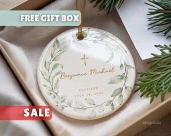 May include: A round, white ceramic ornament with a gold cross and the name "Benjamin Michael" in elegant script. The ornament is decorated with a green and white floral wreath and the text "BAPTIZED JULY 26, 2025". It is presented in a gift box.