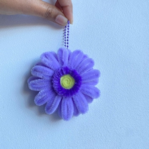 May include: A handmade, plush, purple flower ornament with a yellow center and a purple chain for hanging. The flower has soft, textured petals and a vibrant color palette. The ornament is held by a hand against a light blue background.