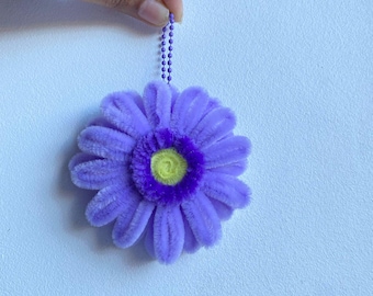 May include: A handmade, plush, purple flower ornament with a yellow center and a purple chain for hanging. The flower has soft, textured petals and a vibrant color palette. The ornament is held by a hand against a light blue background.