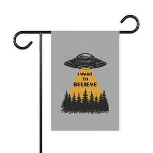 May include: A grey garden flag with a black metal stand. The flag displays a black and white illustration of a UFO above a forest with the text "I WANT TO BELIEVE" in yellow.