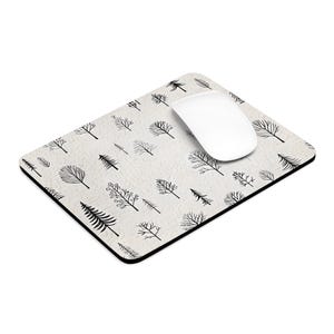 May include: A rectangular mouse mat with a cream background featuring a repeating pattern of black tree and leaf illustrations. A white wireless mouse sits on the mat. The mat has a black border.