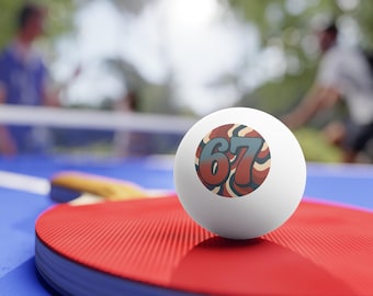 May include: A white table tennis ball featuring the number '67' in a retro design, placed on a red table tennis bat. The bat rests on a blue surface, with a net and blurred figures visible in the background.