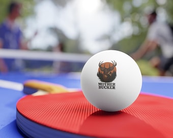 May include: A white ping pong ball with a graphic of a deer head and the text "MOTHER BUCKER". The ball is resting on a red ping pong paddle, with a blue table and net in the background.