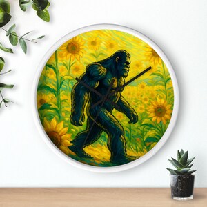 May include: A round wall clock with a white frame. The clock face features a dark green Bigfoot walking through a field of yellow sunflowers. Black clock hands indicate the time.