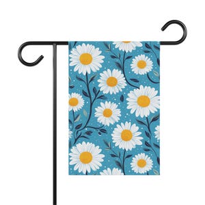 May include: A garden flag featuring a daisy pattern. The flag has a light blue background with white daisies and yellow centers. Dark blue stems and leaves are interspersed among the flowers. The flag is displayed on a black metal stand.