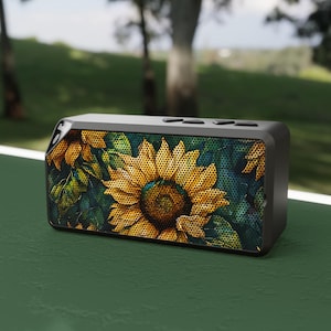 May include: A black portable Bluetooth speaker with a sunflower design. The speaker has a textured surface and a sunflower graphic on the front.