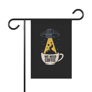 May include: Black garden flag with a humorous design. A UFO shines a beam of light onto a coffee cup, with the text "WE NEED COFFEE". The flag is mounted on a black metal stand with a curved top.