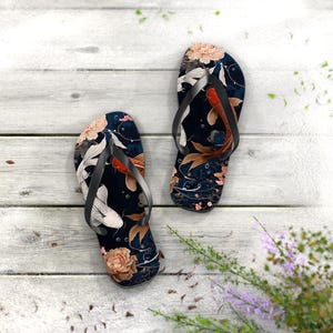 May include: A pair of flip-flops with a dark blue background featuring a koi fish and floral design. The flip-flops have black straps and a white, red, and gold color scheme. The design includes koi fish, flowers, and leaves.