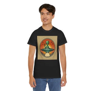 May include: A black t-shirt with a retro-style graphic of a green alien waving from a flying saucer. The graphic is set against a sunset background with stars.