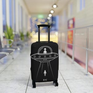 May include: Black rolling suitcase with a white graphic of a UFO, an alien, and a person being beamed up. The word "BYE" is printed at the bottom. The suitcase has a retractable handle and four wheels.