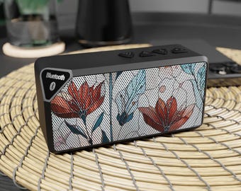 Puede incluir: Un altavoz Bluetooth negro con un diseño floral con flores rojas, azules y blancas. El altavoz tiene una rejilla de malla y un botón que dice "Bluetooth" con un símbolo de Bluetooth.