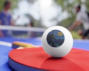 May include: A white ping pong ball featuring a Bigfoot design against a starry night backdrop, with the number 67 on a yellow sign. The ball sits on a red ping pong bat, with a blue table in the foreground.