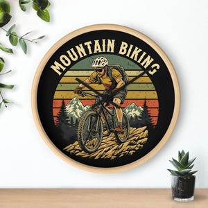 Op de afbeelding: Een ronde houten wandklok met een zwarte wijzerplaat met een vintage-achtige illustratie van een persoon die mountainbike rijdt. De wijzerplaat van de klok heeft de tekst "Mountain Biking" in een witte cirkel met een zonsondergangachtergrond.