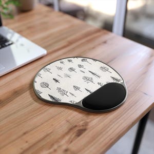 May include: An ergonomic mouse mat with a black wrist rest. The mat features a cream-coloured surface adorned with a repeating pattern of black botanical illustrations. It rests on a wooden desk.