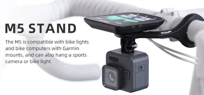 May include: Black M5 stand for a bicycle, holding a sports camera and a cycling computer. The stand is mounted on white handlebars. The text "M5 STAND" is visible.