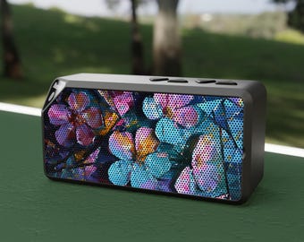 Puede incluir: Un altavoz Bluetooth portátil negro con un diseño floral. El altavoz tiene una rejilla de metal negro y un patrón floral con tonos de azul, rosa y amarillo.