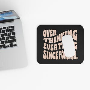 May include: Black rectangular mousepad with the text "OVERTHINKING EVERYTHING SINCE FOREVER" in a retro font. A white computer mouse sits on the mousepad. A laptop is partially visible in the upper left corner.