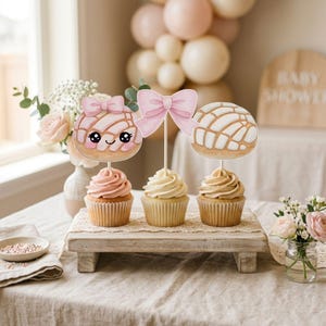 May include: Three cupcakes with decorative toppers, including a pink bow and two concha-shaped designs. The cupcakes sit on a wooden tray, with a baby shower theme in the background. The cupcakes have pink, white, and tan frosting.