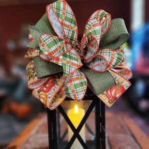 May include: A black metal lantern with a lit white candle inside, topped with a decorative bow. The bow features green and orange plaid ribbon, along with ribbon printed with pumpkins. The lantern sits on a brick surface.