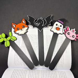 May include: A collection of five whimsical bookmarks. Each bookmark features a different cartoon character: a fox, a bat, a ghost, a balloon animal, and an axolotl. The bookmarks are black with colorful character designs.