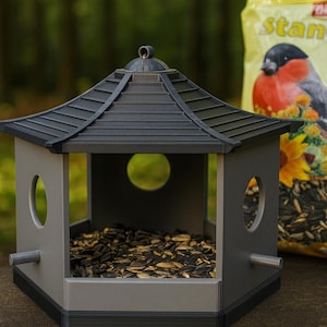 May include: A gray and black pagoda-style bird feeder filled with sunflower seeds. The feeder has a multi-tiered roof and circular openings on the sides. A bag of birdseed with a bird illustration is in the background.
