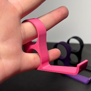 May include: A bright pink finger grip tool is held by a finger. The tool has a ring for the finger and a flat, angled extension. Other tools in black, purple, and other colors are visible in the background. The tools appear to be 3D printed.
