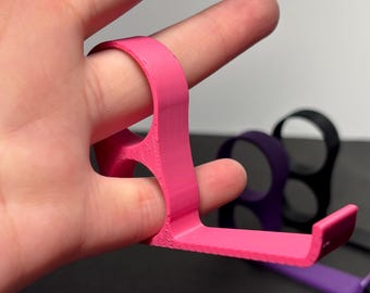 Puede incluir: Una herramienta de agarre para dedos de color rosa brillante es sostenida por un dedo. La herramienta tiene un anillo para el dedo y una extensión plana y en ángulo. Otras herramientas en negro, morado y otros colores son visibles en el fondo. Las herramientas parecen estar impresas en 3D.