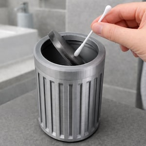 Op de afbeelding: Een cilindrische, zilverkleurige container met verticale groeven, ontworpen voor gebruik in de badkamer. Het deksel is gedeeltelijk open en er wordt een wattenstaafje in geplaatst. De container staat op een grijs oppervlak, wat wijst op een moderne badkamer.