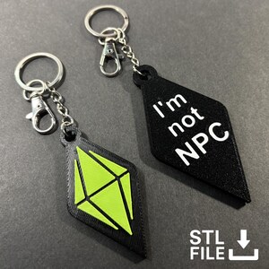 May include: Two black keyrings with silver fixings. One has a green geometric design, and the other says "I'm not NPC" in white. The keyrings are diamond-shaped.