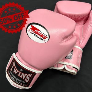 May include: A pair of pink boxing gloves with white trim and the Twins Special logo. The wrist strap features the text Twins Special and a website URL. The gloves are marked as 12 oz. A red circular graphic with the text 50% OFF is visible.