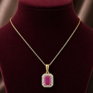 Op de afbeelding: Een gouden ketting met een rechthoekige hanger met een dieproze edelsteen in het midden, omringd door een halo van kleine witte steentjes. De hanger hangt aan een fijne gouden ketting met een gepolijste metalen afwerking.