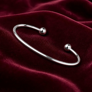 May include: A polished silver-tone cuff bracelet featuring a smooth, open-ended design with a spherical ball bead at each terminal end. The metal surface is highly reflective, showcasing a clean, minimalist aesthetic with a simple, curved band.
