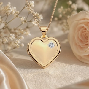 Puede incluir: Un colgante de relicario en forma de corazón de oro suspendido de una delicada cadena de oro. El relicario presenta una superficie lisa y pulida con una pequeña piedra preciosa transparente engastada en estrella en la esquina superior derecha.