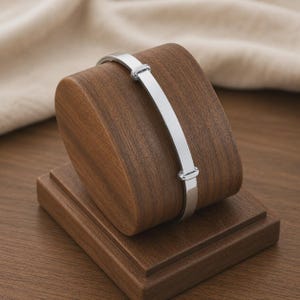 Peut inclure: Un bracelet jonc réglable argenté présenté sur un présentoir à bijoux en bois. Le bracelet a un design rectangulaire avec une fermeture à fermoir. Le présentoir est en bois brun foncé.