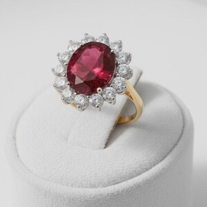 May include: A ring featuring a large, oval-cut, deep red gemstone at the center, surrounded by a halo of twelve round, clear, brilliant-cut stones. The setting is crafted from a gold-toned metal band, providing a classic and elegant appearance.