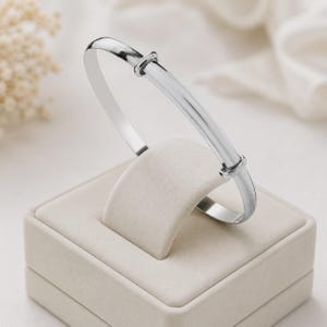 May include: A polished silver adjustable bangle bracelet. The bracelet is presented on a beige jewellery stand. The design is simple and elegant, with a smooth, shiny finish. The adjustable feature allows for a comfortable fit.