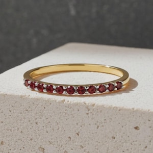 Könnte beinhalten: Ein dünner Goldring mit einer Reihe kleiner, runder, tiefroter Edelsteine, die in silberfarbenen Krappen entlang der vorderen Hälfte des Bandes gefasst sind. Der Ring hat eine polierte Oberfläche und ein schlichtes, schmales Design aus Gold.