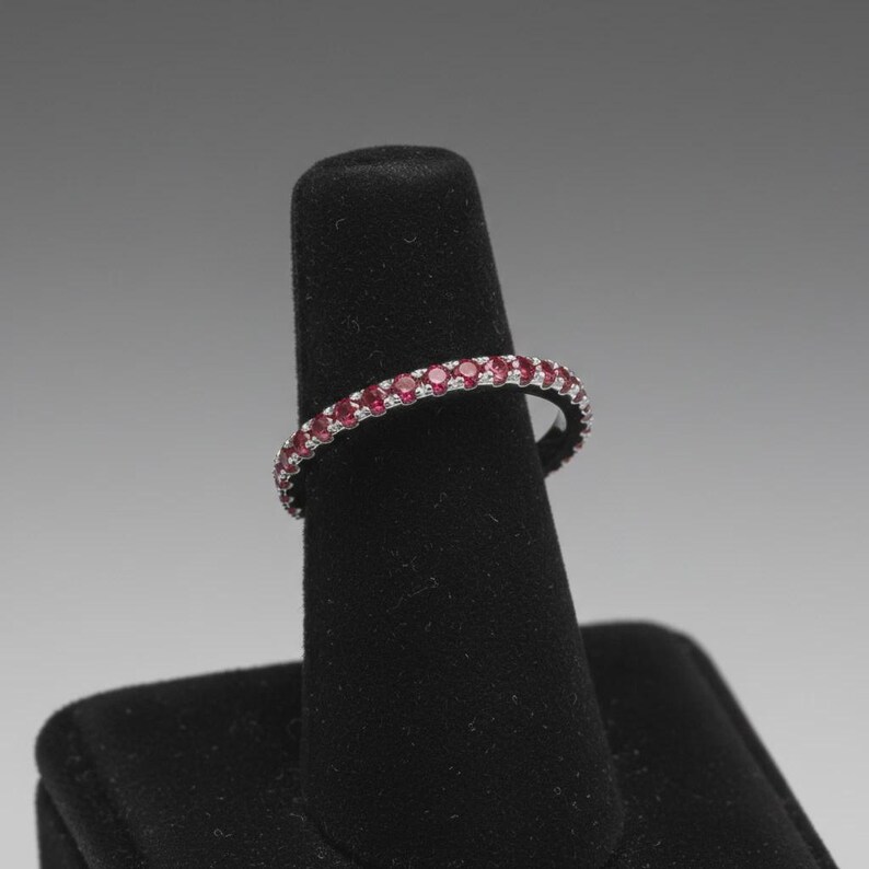 Pode incluir: Um anel de banda fina e circular com uma fila cont&iacute;nua de pequenas pedras preciosas redondas de um vermelho vibrante, cravadas numa arma&ccedil;&atilde;o de metal prateado polido. O anel &eacute; exibido num suporte de joalharia vertical, c&oacute;nico e preto.