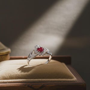 K&ouml;nnte beinhalten: Ein silberner Claddagh-Ring mit einem herzf&ouml;rmigen, roten Edelstein in der Mitte, umgeben von kleineren wei&szlig;en Edelsteinen. Der Ring liegt auf einer beigen Samtoberfl&auml;che in einer Holzbox, wobei Licht und Schatten einen dramatischen Effekt erzeugen.