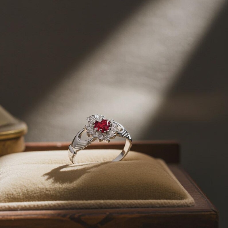 K&ouml;nnte beinhalten: Ein silberner Claddagh-Ring mit einem herzf&ouml;rmigen, roten Edelstein in der Mitte, umgeben von kleineren wei&szlig;en Edelsteinen. Der Ring liegt auf einer beigen Samtoberfl&auml;che in einer Holzbox, wobei Licht und Schatten einen dramatischen Effekt erzeugen.