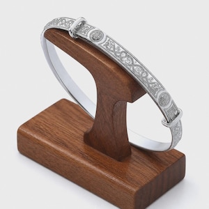May include: A silver bangle bracelet featuring detailed Celtic knot and circular patterns. The bracelet is presented on a wooden stand, highlighting its intricate design. The bracelet's design includes a sun-like emblem. The bracelet is made of silver.