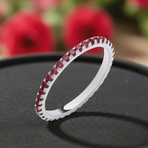 Könnte beinhalten: Ein dünner silberfarbener Ring mit einer durchgehenden Reihe kleiner, runder, dunkelroter Edelsteine, die auf der Außenseite eingefasst sind. Der Ring hat eine polierte metallische Oberfläche und eine gleichmäßige, kreisförmige Form.